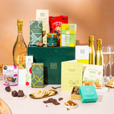 EDEN & CO. Grand Hamper gift box with a full-size Bottega Gold prosecco, dark green gift cylinder, an “Occasion Themed” card, an assortment of chocolate bars, Booja-Booja Chocolate Wonders truffles and Buttermilk snack bars, styled with gold silk against a warm terracotta background.