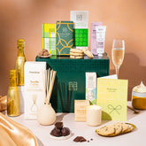 EDEN & CO. Pamper Hamper gift set with a mini Bottega Gold prosecco, dark green gift cylinder, an “Occasion Themed” card, chocolate bars and truffles, a scented candle and Aery hand cream, styled with gold silk against a warm terracotta background.