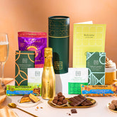 EDEN & CO. plant-based gift hamper with a mini Bottega Gold prosecco, green gift tube, “Occasion Themed” card, chocolate bars in Luscious Orange, White Chocolate Velvet, Signature Chocolate Blend and Salted Caramel, Booja-Booja Chocolate Wonders truffles, Buttermilk Peanut Nougat and Honeycomb Blast bars, chocolate truffles and honeycomb pieces on small plates, and a filled champagne glass, set against a warm terracotta background.