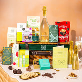 EDEN & CO. Luxury Wicker Hamper filled with a full-size Bottega Gold prosecco, an assortment of chocolate bars, Booja-Booja Chocolate Wonders truffles and Buttermilk snack bars, presented in a dark green trimmed wicker basket with an “Occasion Themed” card, styled against a warm terracotta background.