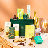EDEN & CO. Food & Drink Hamper with a full-size Bottega Gold prosecco, dark green gift cylinder, an “Occasion Themed” card, an assortment of chocolate bars, Booja-Booja Chocolate Wonders truffles, Buttermilk snack bars, a scented candle and Aery hand cream, styled with gold silk against a warm terracotta background.
