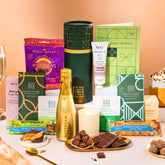 EDEN & CO. plant-based gift hamper with a mini Bottega Gold prosecco, green gift tube, “Occasion Themed” card, chocolate bars in Luscious Orange, White Chocolate Velvet, Signature Chocolate Blend and Salted Caramel, Booja-Booja Chocolate Wonders truffles, Buttermilk Peanut Nougat and Honeycomb Blast bars, chocolate truffles and honeycomb pieces on small plates, and a filled champagne glass, luxury candle and hand cream set against a warm terracotta background.