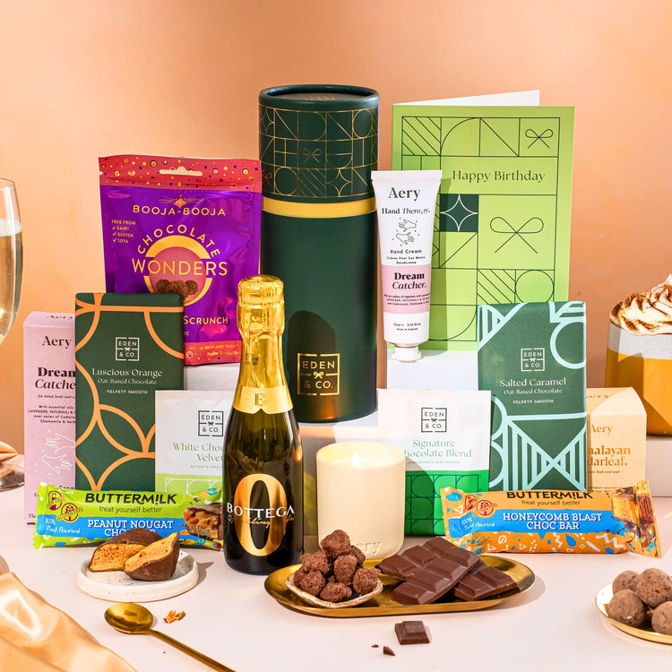 EDEN & CO. plant-based gift hamper with a mini Bottega Zero Black prosecco, green gift tube, “Occasion Themed” card, chocolate bars in Luscious Orange, White Chocolate Velvet, Signature Chocolate Blend and Salted Caramel, Booja-Booja Chocolate Wonders truffles, Buttermilk Peanut Nougat and Honeycomb Blast bars, chocolate truffles and honeycomb pieces on small plates, and a filled champagne glass, luxury candle and hand cream set against a warm terracotta background.