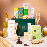 EDEN & CO. Pamper Hamper gift set with a mini Bottega Gold prosecco, dark green gift cylinder, an “Occasion Themed” card, chocolate bars and truffles, a scented candle and Aery hand cream, styled with gold silk against a warm terracotta background.