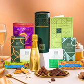 EDEN & CO. plant-based gift hamper with a mini Bottega Gold prosecco, green gift tube, “Occasion Themed” card, chocolate bars in Luscious Orange, White Chocolate Velvet, Signature Chocolate Blend and Salted Caramel, Booja-Booja Chocolate Wonders truffles, Buttermilk Peanut Nougat and Honeycomb Blast bars, chocolate truffles and honeycomb pieces on small plates, and a filled champagne glass, set against a warm terracotta background.