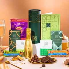 EDEN & CO. plant-based gift hamper with a mini Bottega Gold prosecco, green gift tube, “Occasion Themed” card, chocolate bars in Luscious Orange, White Chocolate Velvet, Signature Chocolate Blend and Salted Caramel, Booja-Booja Chocolate Wonders truffles, Buttermilk Peanut Nougat and Honeycomb Blast bars, chocolate truffles and honeycomb pieces on small plates, and a filled champagne glass, set against a warm terracotta background.