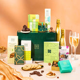 EDEN & CO. Food & Drink Hamper with a full-size Bottega Gold prosecco, dark green gift cylinder, an “Occasion Themed” card, an assortment of chocolate bars, Booja-Booja Chocolate Wonders truffles, Buttermilk snack bars, a scented candle and Aery hand cream, styled with gold silk against a warm terracotta background.