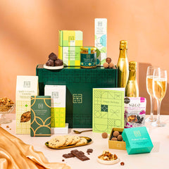 EDEN & CO. Food & Drink Hamper with a full-size Bottega Gold prosecco, dark green gift cylinder, an “Occasion Themed” card, an assortment of chocolate bars, Booja-Booja Chocolate Wonders truffles, Buttermilk snack bars, a scented candle and Aery hand cream, styled with gold silk against a warm terracotta background.