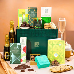 EDEN & CO. Chocolate Hamper letterbox gift with a mini Bottega Zero Black prosecco (0% alcohol-free), green gift tube, an “Occasion Themed” card, an assortment of chocolate bars, Booja-Booja Chocolate Wonders truffles and chocolate truffles on a small plate, styled against a warm terracotta background.