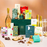 EDEN & CO. Grand Hamper gift box with a full-size Bottega Gold prosecco, dark green gift cylinder, an “Occasion Themed” card, an assortment of chocolate bars, Booja-Booja Chocolate Wonders truffles and Buttermilk snack bars, styled with gold silk against a warm terracotta background.
