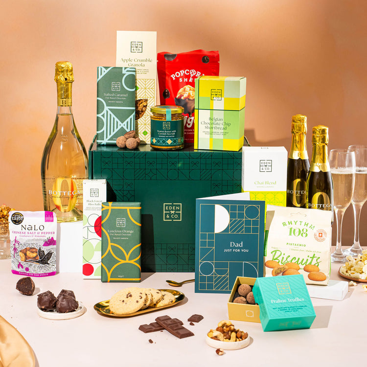 EDEN & CO. Grand Hamper gift box with a full-size Bottega Zero Black prosecco (0% alcohol-free), dark green gift cylinder, an “Occasion Themed” card, an assortment of chocolate bars, Booja-Booja Chocolate Wonders truffles and Buttermilk snack bars, styled with gold silk against a warm terracotta background.