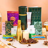EDEN & CO. plant-based gift hamper with a mini Bottega Gold prosecco, green gift tube, “Occasion Themed” card, chocolate bars in Luscious Orange, White Chocolate Velvet, Signature Chocolate Blend and Salted Caramel, Booja-Booja Chocolate Wonders truffles, Buttermilk Peanut Nougat and Honeycomb Blast bars, chocolate truffles and honeycomb pieces on small plates, and a filled champagne glass, luxury candle and hand cream set against a warm terracotta background.