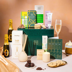 EDEN & CO. Pamper Hamper gift set with a mini Bottega Zero Black prosecco (0% alcohol-free), dark green gift cylinder, an “Occasion Themed” card, chocolate bars and truffles, a scented candle and Aery hand cream, styled with gold silk against a warm terracotta background.
