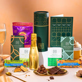 EDEN & CO. plant-based gift hamper with a mini Bottega Gold prosecco, green gift tube, “Occasion Themed” card, chocolate bars in Luscious Orange, White Chocolate Velvet, Signature Chocolate Blend and Salted Caramel, Booja-Booja Chocolate Wonders truffles, Buttermilk Peanut Nougat and Honeycomb Blast bars, chocolate truffles and honeycomb pieces on small plates, and a filled champagne glass, set against a warm terracotta background.