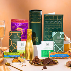 EDEN & CO. plant-based gift hamper with a mini Bottega Gold prosecco, green gift tube, “Occasion Themed” card, chocolate bars in Luscious Orange, White Chocolate Velvet, Signature Chocolate Blend and Salted Caramel, Booja-Booja Chocolate Wonders truffles, Buttermilk Peanut Nougat and Honeycomb Blast bars, chocolate truffles and honeycomb pieces on small plates, and a filled champagne glass, set against a warm terracotta background.