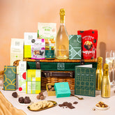 EDEN & CO. Luxury Wicker Hamper filled with a full-size Bottega Gold prosecco, an assortment of chocolate bars, Booja-Booja Chocolate Wonders truffles and Buttermilk snack bars, presented in a dark green trimmed wicker basket with an “Occasion Themed” card, styled against a warm terracotta background.