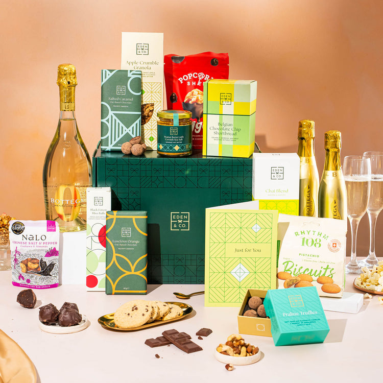 EDEN & CO. Grand Hamper gift box with a full-size Bottega Gold prosecco, dark green gift cylinder, an “Occasion Themed” card, an assortment of chocolate bars, Booja-Booja Chocolate Wonders truffles and Buttermilk snack bars, styled with gold silk against a warm terracotta background.