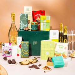 EDEN & CO. Grand Hamper gift box with a full-size Bottega Zero Black prosecco (0% alcohol-free), dark green gift cylinder, an “Occasion Themed” card, an assortment of chocolate bars, Booja-Booja Chocolate Wonders truffles and Buttermilk snack bars, styled with gold silk against a warm terracotta background.