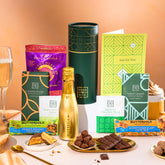 EDEN & CO. plant-based gift hamper with a mini Bottega Gold prosecco, green gift tube, “Occasion Themed” card, chocolate bars in Luscious Orange, White Chocolate Velvet, Signature Chocolate Blend and Salted Caramel, Booja-Booja Chocolate Wonders truffles, Buttermilk Peanut Nougat and Honeycomb Blast bars, chocolate truffles and honeycomb pieces on small plates, and a filled champagne glass, set against a warm terracotta background.