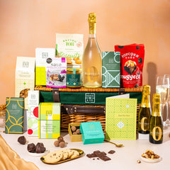 EDEN & CO. Luxury Wicker Hamper filled with a full-size Bottega Zero Black prosecco (0% alcohol-free), an assortment of chocolate bars, Booja-Booja Chocolate Wonders truffles and Buttermilk snack bars, presented in a dark green trimmed wicker basket with an “Occasion Themed” card, styled against a warm terracotta background.