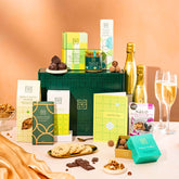 EDEN & CO. Food & Drink Hamper with a full-size Bottega Gold prosecco, dark green gift cylinder, an “Occasion Themed” card, an assortment of chocolate bars, Booja-Booja Chocolate Wonders truffles, Buttermilk snack bars, a scented candle and Aery hand cream, styled with gold silk against a warm terracotta background.