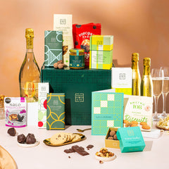 EDEN & CO. Grand Hamper gift box with a full-size Bottega Gold prosecco, dark green gift cylinder, an “Occasion Themed” card, an assortment of chocolate bars, Booja-Booja Chocolate Wonders truffles and Buttermilk snack bars, styled with gold silk against a warm terracotta background.