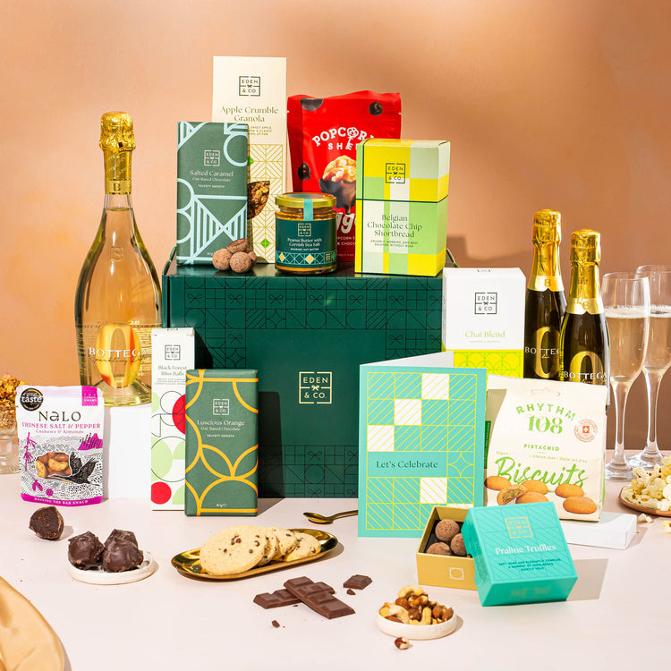 EDEN & CO. Grand Hamper gift box with a full-size Bottega Zero Black prosecco (0% alcohol-free), dark green gift cylinder, an “Occasion Themed” card, an assortment of chocolate bars, Booja-Booja Chocolate Wonders truffles and Buttermilk snack bars, styled with gold silk against a warm terracotta background.