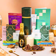 EDEN & CO. plant-based gift hamper with a mini Bottega Zero Black prosecco, green gift tube, “Occasion Themed” card, chocolate bars in Luscious Orange, White Chocolate Velvet, Signature Chocolate Blend and Salted Caramel, Booja-Booja Chocolate Wonders truffles, Buttermilk Peanut Nougat and Honeycomb Blast bars, chocolate truffles and honeycomb pieces on small plates, and a filled champagne glass, luxury candle and hand cream set against a warm terracotta background.