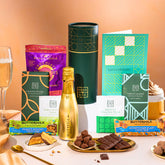 EDEN & CO. plant-based gift hamper with a mini Bottega Gold prosecco, green gift tube, “Occasion Themed” card, chocolate bars in Luscious Orange, White Chocolate Velvet, Signature Chocolate Blend and Salted Caramel, Booja-Booja Chocolate Wonders truffles, Buttermilk Peanut Nougat and Honeycomb Blast bars, chocolate truffles and honeycomb pieces on small plates, and a filled champagne glass, set against a warm terracotta background.
