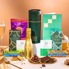 EDEN & CO. plant-based gift hamper with a mini Bottega Gold prosecco, green gift tube, “Occasion Themed” card, chocolate bars in Luscious Orange, White Chocolate Velvet, Signature Chocolate Blend and Salted Caramel, Booja-Booja Chocolate Wonders truffles, Buttermilk Peanut Nougat and Honeycomb Blast bars, chocolate truffles and honeycomb pieces on small plates, and a filled champagne glass, set against a warm terracotta background.