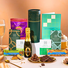 EDEN & CO. plant-based gift hamper with a mini Bottega Zero Black prosecco, green gift tube, “Occasion Themed” card, chocolate bars in Luscious Orange, White Chocolate Velvet, Signature Chocolate Blend and Salted Caramel, Booja-Booja Chocolate Wonders truffles, Buttermilk Peanut Nougat and Honeycomb Blast bars, chocolate truffles and honeycomb pieces on small plates, and a filled champagne glass, set against a warm terracotta background.