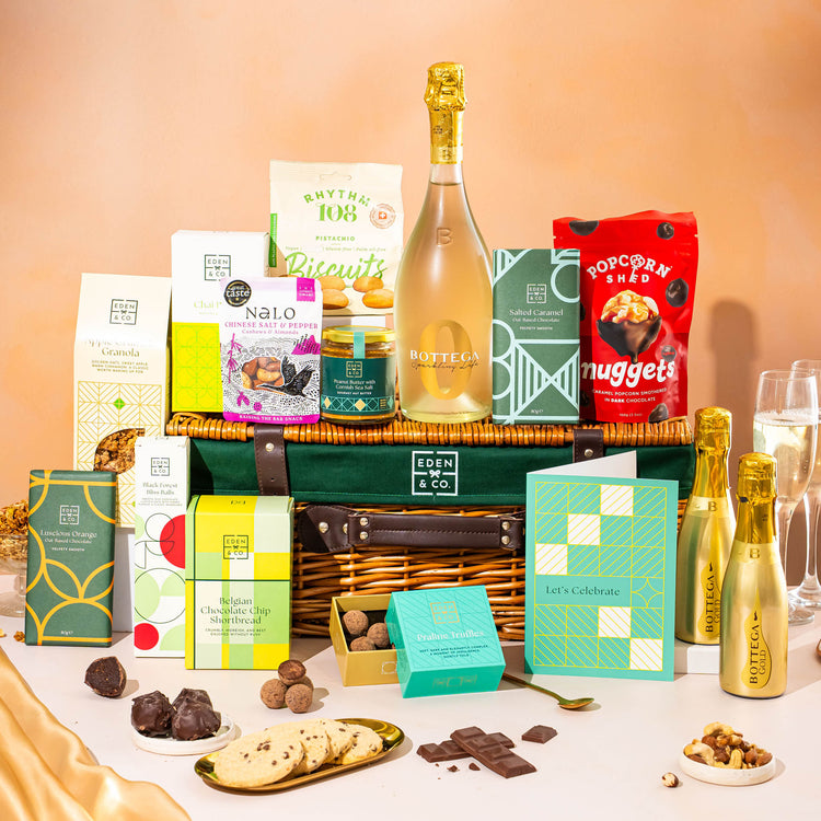 EDEN & CO. Luxury Wicker Hamper filled with a full-size Bottega Gold prosecco, an assortment of chocolate bars, Booja-Booja Chocolate Wonders truffles and Buttermilk snack bars, presented in a dark green trimmed wicker basket with an “Occasion Themed” card, styled against a warm terracotta background.