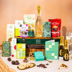EDEN & CO. Luxury Wicker Hamper filled with a full-size Bottega Zero Black prosecco (0% alcohol-free), an assortment of chocolate bars, Booja-Booja Chocolate Wonders truffles and Buttermilk snack bars, presented in a dark green trimmed wicker basket with an “Occasion Themed” card, styled against a warm terracotta background.