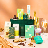 EDEN & CO. Food & Drink Hamper with a full-size Bottega Gold prosecco, dark green gift cylinder, an “Occasion Themed” card, an assortment of chocolate bars, Booja-Booja Chocolate Wonders truffles, Buttermilk snack bars, a scented candle and Aery hand cream, styled with gold silk against a warm terracotta background.