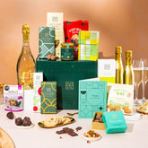 EDEN & CO. Grand Hamper gift box with a full-size Bottega Gold prosecco, dark green gift cylinder, an “Occasion Themed” card, an assortment of chocolate bars, Booja-Booja Chocolate Wonders truffles and Buttermilk snack bars, styled with gold silk against a warm terracotta background.