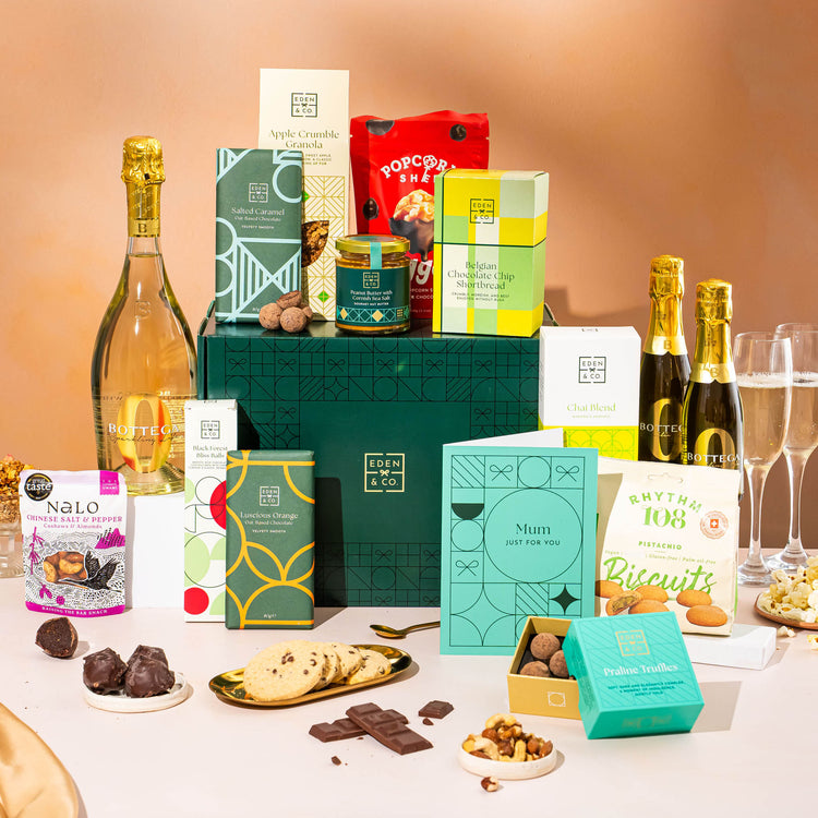 EDEN & CO. Grand Hamper gift box with a full-size Bottega Zero Black prosecco (0% alcohol-free), dark green gift cylinder, an “Occasion Themed” card, an assortment of chocolate bars, Booja-Booja Chocolate Wonders truffles and Buttermilk snack bars, styled with gold silk against a warm terracotta background.