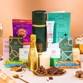 EDEN & CO. plant-based gift hamper with a mini Bottega Gold prosecco, green gift tube, “Occasion Themed” card, chocolate bars in Luscious Orange, White Chocolate Velvet, Signature Chocolate Blend and Salted Caramel, Booja-Booja Chocolate Wonders truffles, Buttermilk Peanut Nougat and Honeycomb Blast bars, chocolate truffles and honeycomb pieces on small plates, and a filled champagne glass, luxury candle and hand cream set against a warm terracotta background.