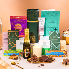 EDEN & CO. plant-based gift hamper with a mini Bottega Zero Black prosecco, green gift tube, “Occasion Themed” card, chocolate bars in Luscious Orange, White Chocolate Velvet, Signature Chocolate Blend and Salted Caramel, Booja-Booja Chocolate Wonders truffles, Buttermilk Peanut Nougat and Honeycomb Blast bars, chocolate truffles and honeycomb pieces on small plates, and a filled champagne glass, luxury candle and hand cream set against a warm terracotta background.