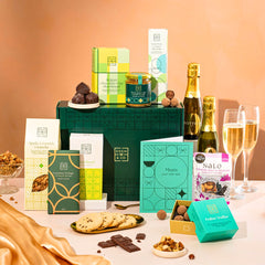 EDEN & CO. Food & Drink Hamper with a full-size Bottega Zero Black prosecco (0% alcohol-free), dark green gift cylinder, an “Occasion Themed” card, an assortment of chocolate bars, Booja-Booja Chocolate Wonders truffles, Buttermilk snack bars, a scented candle and Aery hand cream, styled with gold silk against a warm terracotta background.