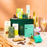 EDEN & CO. Food & Drink Hamper with a full-size Bottega Gold prosecco, dark green gift cylinder, an “Occasion Themed” card, an assortment of chocolate bars, Booja-Booja Chocolate Wonders truffles, Buttermilk snack bars, a scented candle and Aery hand cream, styled with gold silk against a warm terracotta background.