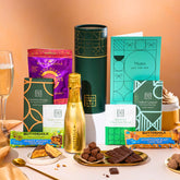 EDEN & CO. plant-based gift hamper with a mini Bottega Gold prosecco, green gift tube, “Occasion Themed” card, chocolate bars in Luscious Orange, White Chocolate Velvet, Signature Chocolate Blend and Salted Caramel, Booja-Booja Chocolate Wonders truffles, Buttermilk Peanut Nougat and Honeycomb Blast bars, chocolate truffles and honeycomb pieces on small plates, and a filled champagne glass, set against a warm terracotta background.