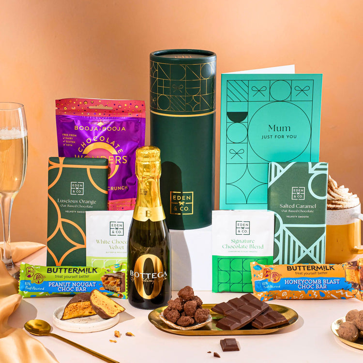 EDEN & CO. plant-based gift hamper with a mini Bottega Zero Black prosecco, green gift tube, “Occasion Themed” card, chocolate bars in Luscious Orange, White Chocolate Velvet, Signature Chocolate Blend and Salted Caramel, Booja-Booja Chocolate Wonders truffles, Buttermilk Peanut Nougat and Honeycomb Blast bars, chocolate truffles and honeycomb pieces on small plates, and a filled champagne glass, set against a warm terracotta background.