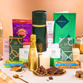 EDEN & CO. plant-based gift hamper with a mini Bottega Gold prosecco, green gift tube, “Occasion Themed” card, chocolate bars in Luscious Orange, White Chocolate Velvet, Signature Chocolate Blend and Salted Caramel, Booja-Booja Chocolate Wonders truffles, Buttermilk Peanut Nougat and Honeycomb Blast bars, chocolate truffles and honeycomb pieces on small plates, and a filled champagne glass, luxury candle and hand cream set against a warm terracotta background.