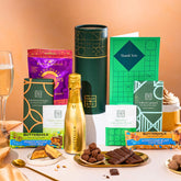 EDEN & CO. plant-based gift hamper with a mini Bottega Gold prosecco, green gift tube, “Occasion Themed” card, chocolate bars in Luscious Orange, White Chocolate Velvet, Signature Chocolate Blend and Salted Caramel, Booja-Booja Chocolate Wonders truffles, Buttermilk Peanut Nougat and Honeycomb Blast bars, chocolate truffles and honeycomb pieces on small plates, and a filled champagne glass, set against a warm terracotta background.