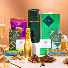 EDEN & CO. plant-based gift hamper with a mini Bottega Gold prosecco, green gift tube, “Occasion Themed” card, chocolate bars in Luscious Orange, White Chocolate Velvet, Signature Chocolate Blend and Salted Caramel, Booja-Booja Chocolate Wonders truffles, Buttermilk Peanut Nougat and Honeycomb Blast bars, chocolate truffles and honeycomb pieces on small plates, and a filled champagne glass, set against a warm terracotta background.