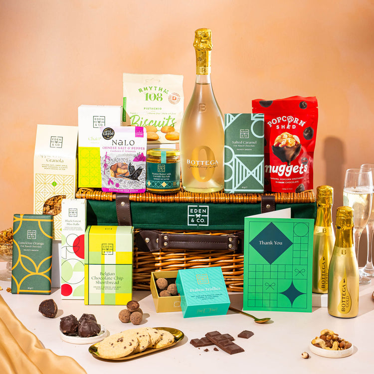 EDEN & CO. Luxury Wicker Hamper filled with a full-size Bottega Gold prosecco, an assortment of chocolate bars, Booja-Booja Chocolate Wonders truffles and Buttermilk snack bars, presented in a dark green trimmed wicker basket with an “Occasion Themed” card, styled against a warm terracotta background.