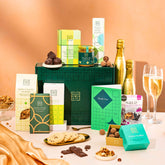 EDEN & CO. Food & Drink Hamper with a full-size Bottega Gold prosecco, dark green gift cylinder, an “Occasion Themed” card, an assortment of chocolate bars, Booja-Booja Chocolate Wonders truffles, Buttermilk snack bars, a scented candle and Aery hand cream, styled with gold silk against a warm terracotta background.