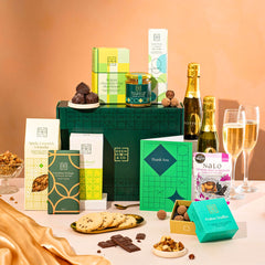 EDEN & CO. Food & Drink Hamper with a full-size Bottega Zero Black prosecco (0% alcohol-free), dark green gift cylinder, an “Occasion Themed” card, an assortment of chocolate bars, Booja-Booja Chocolate Wonders truffles, Buttermilk snack bars, a scented candle and Aery hand cream, styled with gold silk against a warm terracotta background.