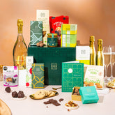 EDEN & CO. Grand Hamper gift box with a full-size Bottega Gold prosecco, dark green gift cylinder, an “Occasion Themed” card, an assortment of chocolate bars, Booja-Booja Chocolate Wonders truffles and Buttermilk snack bars, styled with gold silk against a warm terracotta background.