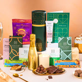 EDEN & CO. plant-based gift hamper with a mini Bottega Gold prosecco, green gift tube, “Occasion Themed” card, chocolate bars in Luscious Orange, White Chocolate Velvet, Signature Chocolate Blend and Salted Caramel, Booja-Booja Chocolate Wonders truffles, Buttermilk Peanut Nougat and Honeycomb Blast bars, chocolate truffles and honeycomb pieces on small plates, and a filled champagne glass, luxury candle and hand cream set against a warm terracotta background.